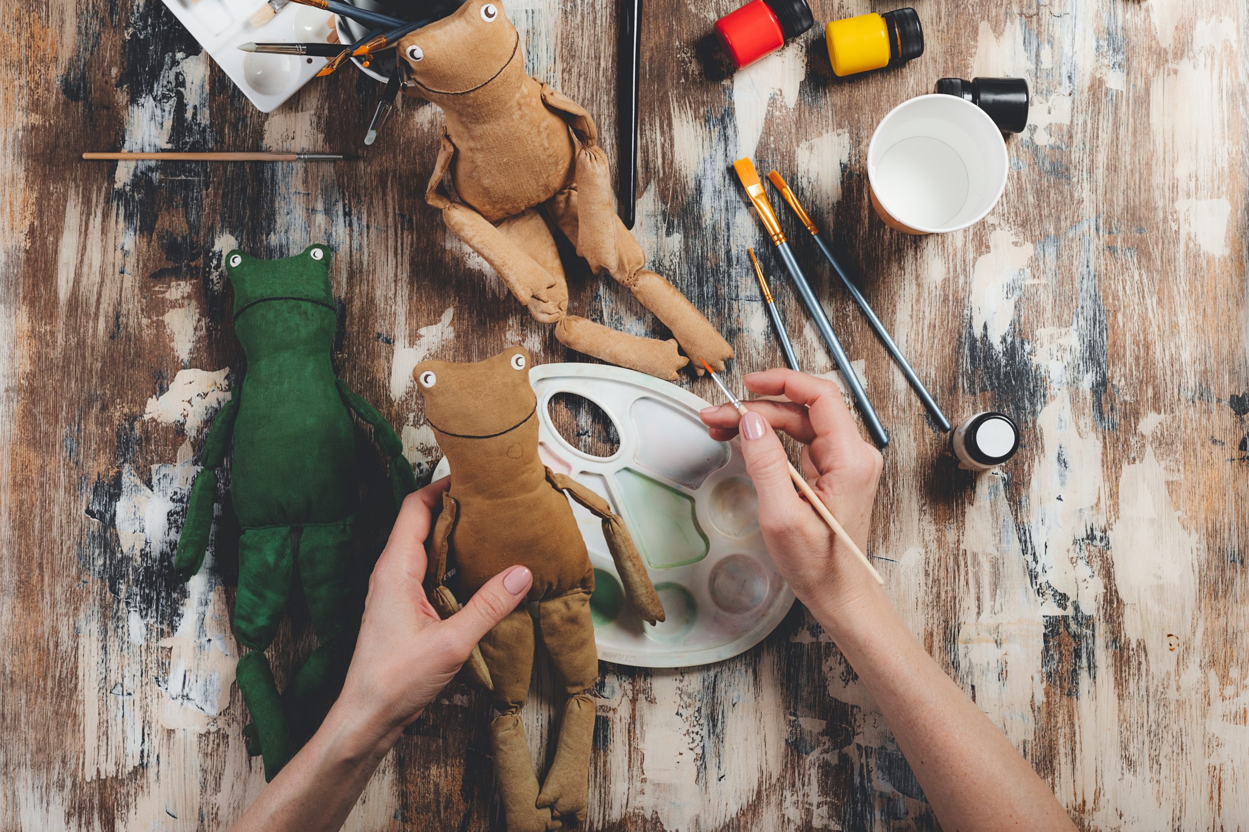 Handmade soft Doll frog. DIY gift making. Human hands hold frog soft toy. Needlework accessories on grey background. Handmade house decor. Process of crafting a vintage soft toy. Top view